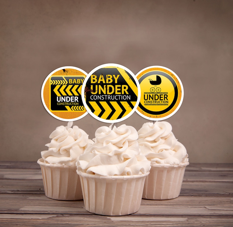 Puede incluir: Tres adornos para cupcakes amarillos y negros con el texto "Baby Under Construction" y un icono de cochecito. Los adornos est&aacute;n encima de cupcakes con glaseado blanco.