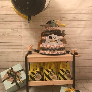 Puede incluir: Un pastel de pa&ntilde;ales para un baby shower con tem&aacute;tica de construcci&oacute;n con un globo negro y amarillo, un letrero que dice "&iexcl;Es un ni&ntilde;o!" y peque&ntilde;as bolsas de regalo amarillas con dise&ntilde;os de cinta de advertencia negra y amarilla.