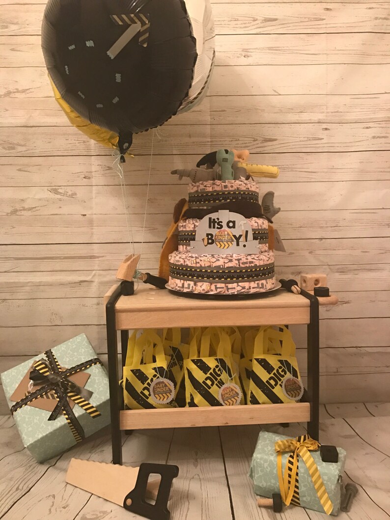 Puede incluir: Un pastel de pa&ntilde;ales para un baby shower con tem&aacute;tica de construcci&oacute;n con un globo negro y amarillo, un letrero que dice "&iexcl;Es un ni&ntilde;o!" y peque&ntilde;as bolsas de regalo amarillas con dise&ntilde;os de cinta de advertencia negra y amarilla.