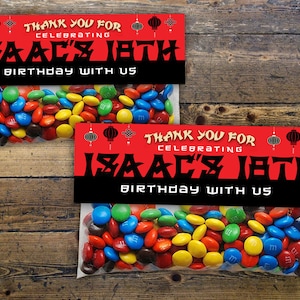Peut inclure: Un sac de fête rouge et noir avec une bannière rouge et noire qui dit "Merci d'avoir célébré le 18e anniversaire d'Isaac avec nous". Le sac est rempli de bonbons M&M's colorés.