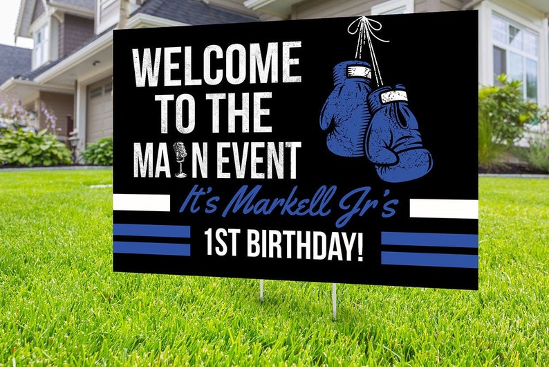 May include: Black and white yard sign with blue boxing gloves hanging from the top. The sign reads "Welcome to the Main Event It's Markell Jr's 1st Birthday!"