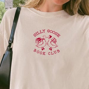 May include: Ivory-colored t-shirt with the text "SILLY GOOSE BOOK CLUB" in red. The graphic features two geese with books. The shirt is a relaxed fit.