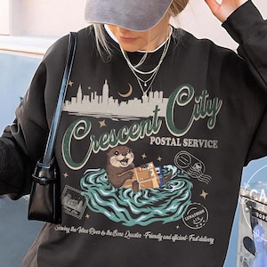 May include: A dark gray sweatshirt with a graphic featuring an otter delivering mail in a river, with the text "Crescent City Postal Service." The design includes a cityscape, moon, and the phrase "Serving the River to the Bone Quarter." A gray baseball cap is worn.