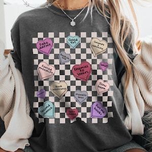 May include: Dark gray sweatshirt with a checkerboard pattern and heart-shaped graphics. The hearts feature phrases like "Fake Dating," "Enemies to Lovers," and "Buy Me Books." The word "Pepper" is printed on the bottom right.