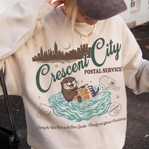 Op de afbeelding: Een zandkleurig sweatshirt met een afbeelding van een otter, de woorden "Crescent City Postal Service" en een stadsgezicht. Het sweatshirt wordt gecombineerd met een bruine baseballcap met de tekst "La Paris".