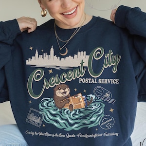 May include: Navy blue sweatshirt with a graphic featuring an otter delivering mail in a river, with the text "Crescent City Postal Service." The design includes a cityscape and the words "Serving the Vieux River & the Bone Quarter."