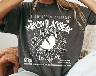 May include: A charcoal gray short-sleeve t-shirt featuring a white graphic of a large reptilian eye surrounded by scales. The text reads THE THIRTEEN PRESENTS MANON BLACKBEAK WORLD TOUR FEATURING ASTERIN SORREL VESTA FALINE FALLON EDDA BRIAR THEA KAYA LINNEA GAVRIEL.
