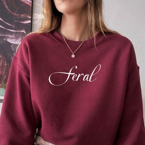 K&ouml;nnte beinhalten: Ein maroonfarbener Pullover mit dem Wort "Feral" in wei&szlig;er Kursivschrift auf der Vorderseite. Der Pullover wird von einer Person getragen, die einen karierten Rock und schwarze Strumpfhosen tr&auml;gt.
