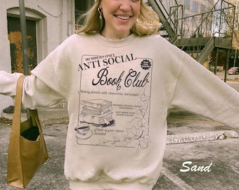 May include: A beige sweatshirt with a graphic print of a book club logo. The logo says "Members Only Anti Social Book Club" and includes a stack of books, a cup of coffee, and leaves. The text below the logo says "Making friends with characters, not people!" and "Drink of your choice included."
