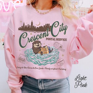 Op de afbeelding: Lichtroze sweatshirt met een afbeelding van een stadsgezicht, de tekst "Crescent City Postal Service" en een otter met pakketten. De tekst "Sewing the Vatros River & the Bone Quarter" is ook zichtbaar.