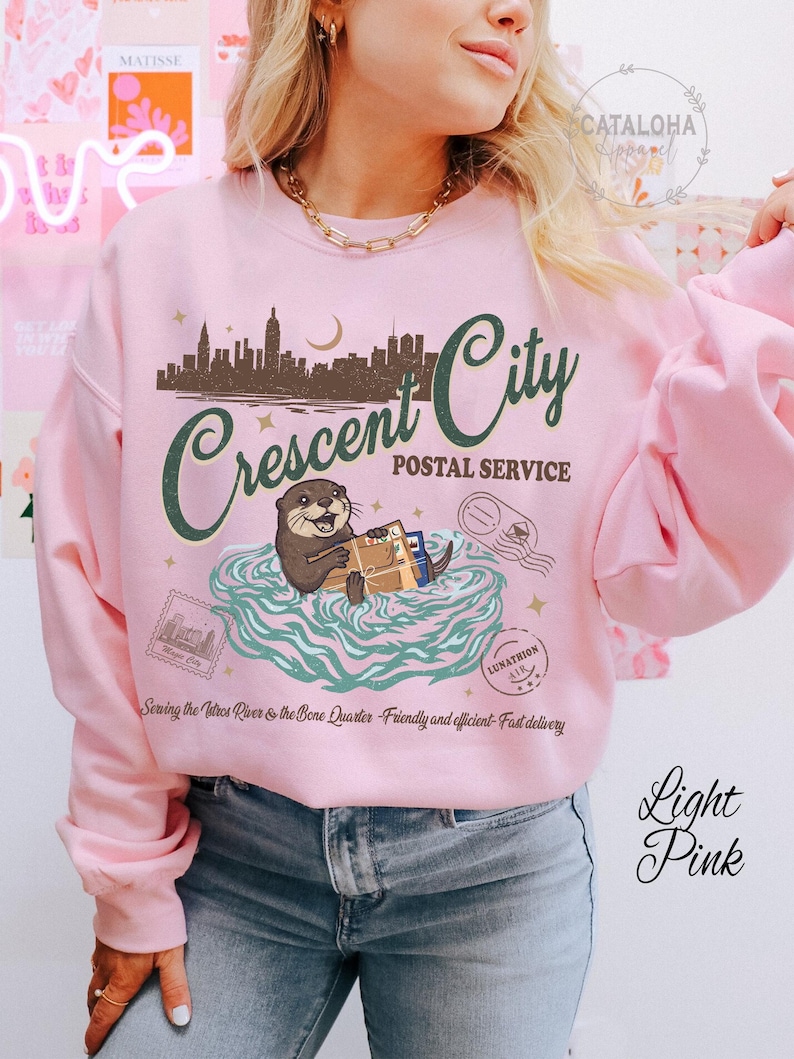 Op de afbeelding: Lichtroze sweatshirt met een afbeelding van een stadsgezicht, de tekst "Crescent City Postal Service" en een otter met pakketten. De tekst "Sewing the Vatros River & the Bone Quarter" is ook zichtbaar.