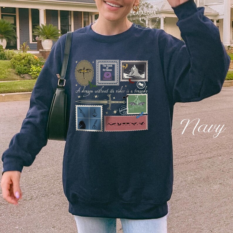 Puede incluir: Sudadera azul marino con un dise&ntilde;o gr&aacute;fico que presenta ilustraciones estilo sello postal y el texto "A dragon without its rider is a tragedy". El dise&ntilde;o incluye una espada, estrellas e im&aacute;genes de fantas&iacute;a.