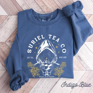 May include: Indigo blue sweatshirt with the text "SURIEL TEA CO" and a graphic of a hooded figure holding a cup. The design includes the phrases "HOT TEA" and "HIGH LORD", with yellow roses and stars. The text "Hottest tea in Prythion" is also present.