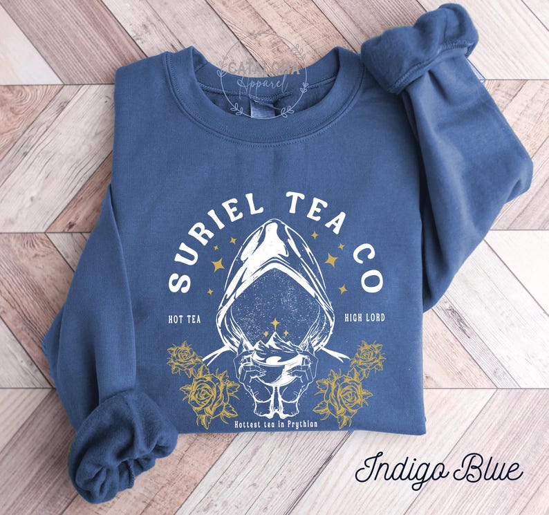 May include: Indigo blue sweatshirt with the text "SURIEL TEA CO" and a graphic of a hooded figure holding a cup. The design includes the phrases "HOT TEA" and "HIGH LORD", with yellow roses and stars. The text "Hottest tea in Prythion" is also present.