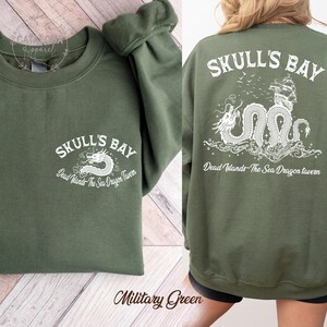 May include: Military green sweatshirt with white text and graphic. The front features "SKULL'S BAY" and a dragon design. The back displays the same text and a detailed dragon and ship illustration. The text "Dead Islands The Sea Dragon Tavern" is also present.