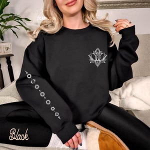 May include: Black sweatshirt with a white lotus flower design on the chest and a series of white circular and dot patterns on the left sleeve. The word "Black" is printed in cursive on the lower left.