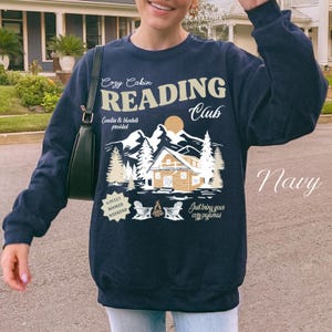 May include: Navy blue sweatshirt featuring a graphic for a "Cozy Cabin Reading Club." The design includes a cabin, mountains, trees, and the text "Just bring your cosy pyjamas." The sweatshirt also has the text "A Fully Booked Weekend."