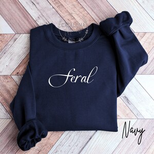 K&ouml;nnte beinhalten: Marineblauer Sweatshirt mit dem Wort "Feral" in wei&szlig;er Kursivschrift.