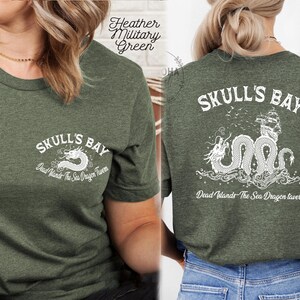 May include: Heather military green t-shirt with the text "Skull's Bay" and a white dragon graphic on the front. The back of the shirt features a larger graphic of a sea dragon, ship, and the text "Skull's Bay" and "Dead Winds - The Sea Dragon Tavern."