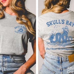 May include: Athletic grey t-shirt with blue graphic text and a sea dragon design. The front of the shirt reads "Skull's Bay" with a sea dragon logo. The back of the shirt features a larger sea dragon and ship design with the text "Skull's Bay" and "Dead Islands The Sea Dragon Inn".
