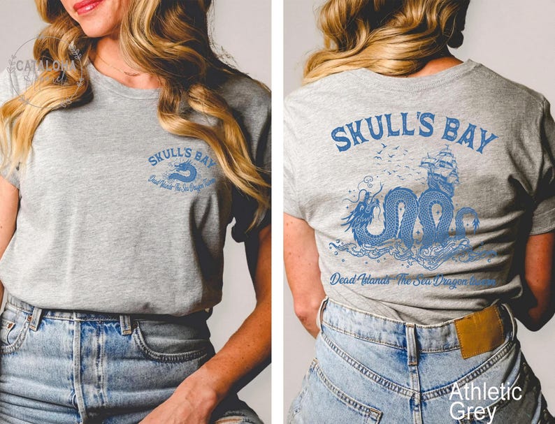 May include: Athletic grey t-shirt with blue graphic text and a sea dragon design. The front of the shirt reads "Skull's Bay" with a sea dragon logo. The back of the shirt features a larger sea dragon and ship design with the text "Skull's Bay" and "Dead Islands The Sea Dragon Inn".