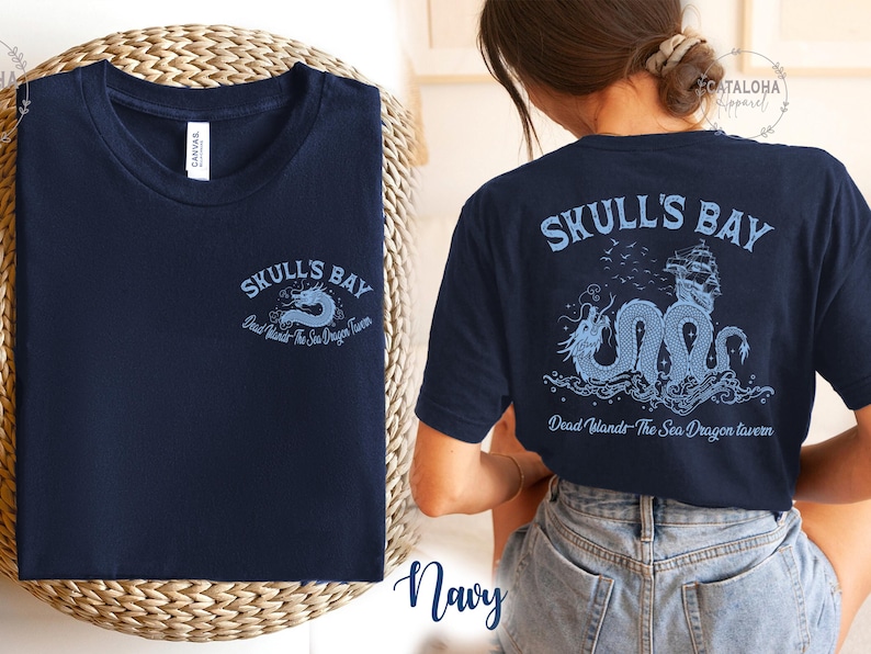 May include: Navy blue t-shirt with "SKULL'S BAY" text and a light blue dragon illustration. The text "Dead Islands The Sea Dragon Tavern" is below the dragon. The shirt is displayed on a woven surface.