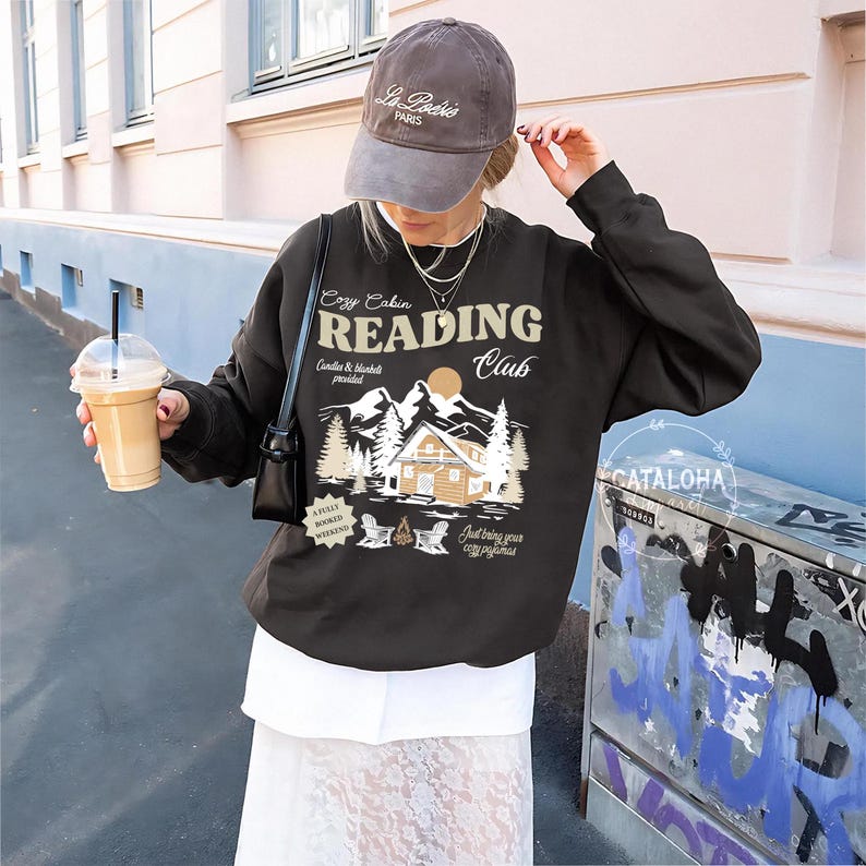 May include: A dark grey sweatshirt with a graphic that reads "Cozy Cabin Reading Club." The graphic features a cabin, mountains, and trees. The sweatshirt also has the text "Candle & blanket provided" and "Just bring your cozy pyjamas."