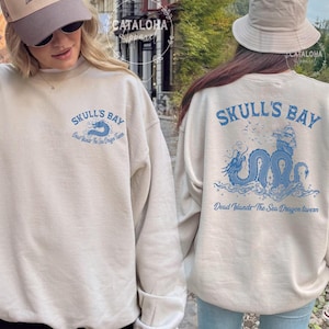 May include: Sand-colored sweatshirt with blue graphic text and a sea dragon design. The front of the sweatshirt reads "Skull's Bay" with a sea creature illustration. The back features a larger design with a ship and the text "Dead Islands The Sea Dragon Tavern."