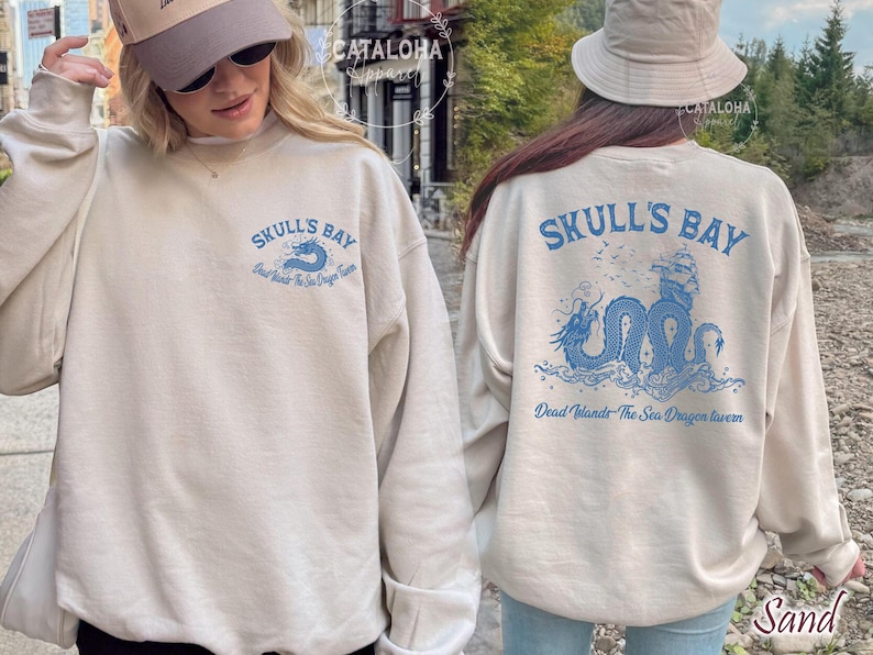 May include: Sand-colored sweatshirt with blue graphic text and a sea dragon design. The front of the sweatshirt reads "Skull's Bay" with a sea creature illustration. The back features a larger design with a ship and the text "Dead Islands The Sea Dragon Tavern."