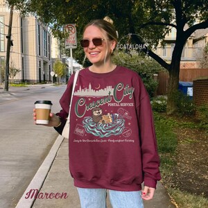 Op de afbeelding: Bordeaux sweatshirt met een afbeelding met de tekst "Crescent City Postal Service". Het ontwerp toont een otter die pakketten vasthoudt in een waterlandschap, met daarboven een silhouet van een stadsgezicht. De tekst "Serving the New Orleans to the New Orleans Quarter. Friendly and efficient fast delivery" is ook opgenomen.
