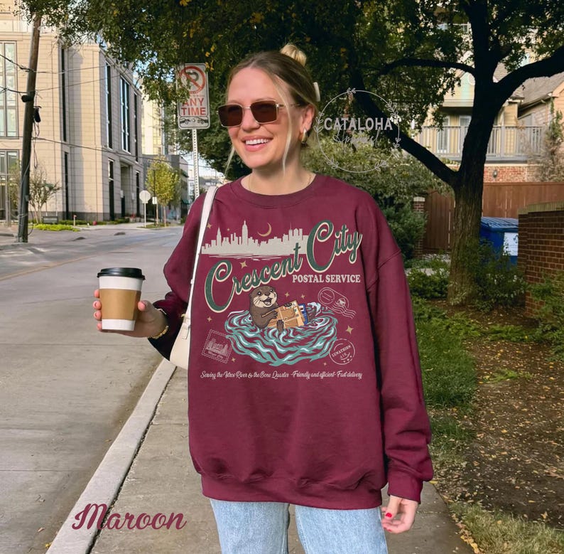 Op de afbeelding: Bordeaux sweatshirt met een afbeelding met de tekst "Crescent City Postal Service". Het ontwerp toont een otter die pakketten vasthoudt in een waterlandschap, met daarboven een silhouet van een stadsgezicht. De tekst "Serving the New Orleans to the New Orleans Quarter. Friendly and efficient fast delivery" is ook opgenomen.