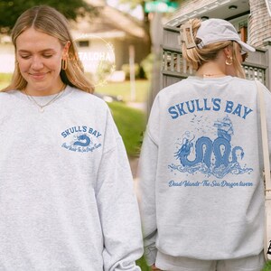 May include: Light gray sweatshirt with the text "SKULL'S BAY" in blue on the front and back. The back features a blue illustration of a sea dragon, ship, and text "Dead Islands The Sea Dragon Tavern".