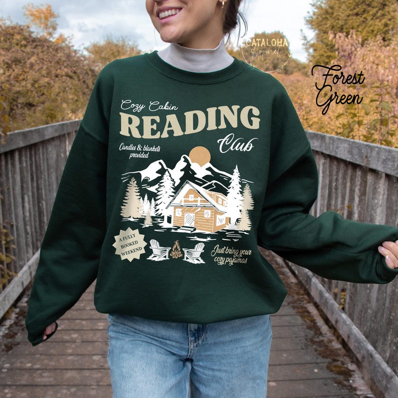May include: Dark green sweatshirt featuring the text "Cozy Cabin Reading Club" with a cabin, mountains, and trees graphic. The design includes the phrases "Candles & blankets provided" and "Just bring your cozy pajamas."