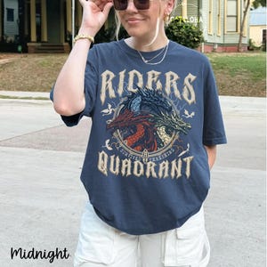 May include: Midnight blue t-shirt featuring a circular graphic with three dragons in red, blue, and green. The text on the shirt reads "RIDERS" above the graphic and "QUADRANT" below. The shirt is worn with white pants.