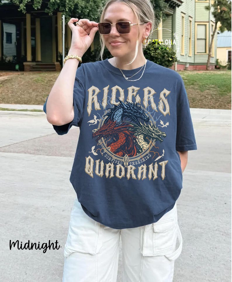 May include: Midnight blue t-shirt featuring a circular graphic with three dragons in red, blue, and green. The text on the shirt reads "RIDERS" above the graphic and "QUADRANT" below. The shirt is worn with white pants.