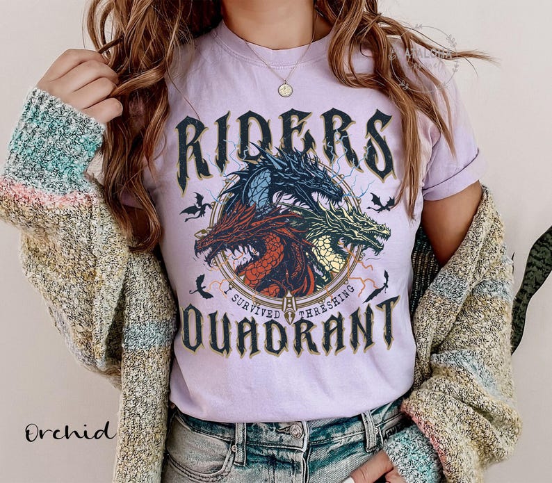 May include: A light purple t-shirt with a graphic featuring three dragons in red, blue, and green, with the words "RIDERS QUADRANT" and "I SURVIVED THRASHING" in a gothic font. The shirt is worn with a multi-colored cardigan.