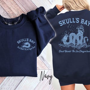 May include: Navy blue sweatshirt with "SKULL'S BAY" text and a dragon graphic on the front and back. The back design includes a ship and the text "Dead Islands - The Sea Dragon Tavern". The sweatshirt is a crew neck style.