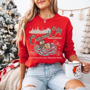 Op de afbeelding: Een rode sweatshirt met een afbeelding van een otter die pakketten bezorgt, met de tekst "Crescent City Postal Service". Het ontwerp bevat een silhouet van een stadsgezicht en postelementen. Een witte mok met een afbeelding van de kerstman wordt in de hand gehouden.