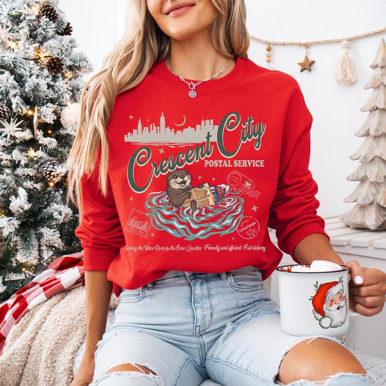 Op de afbeelding: Een rode sweatshirt met een afbeelding van een otter die pakketten bezorgt, met de tekst "Crescent City Postal Service". Het ontwerp bevat een silhouet van een stadsgezicht en postelementen. Een witte mok met een afbeelding van de kerstman wordt in de hand gehouden.