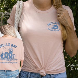 May include: Heather peach-colored t-shirt with a crew neck. The front of the shirt has a small blue dragon graphic and the words "Skull's Bay". The back of the shirt features a larger blue dragon design with the text "Dead Islands The Sea Dragon Swims".