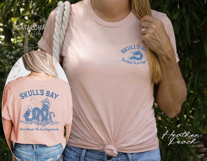 May include: Heather peach-colored t-shirt with a crew neck. The front of the shirt has a small blue dragon graphic and the words "Skull's Bay". The back of the shirt features a larger blue dragon design with the text "Dead Islands The Sea Dragon Swims".