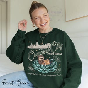 Op de afbeelding: Bost groene sweatshirt met een afbeelding van een otter die post bezorgt voor Crescent City Postal Service. Het ontwerp bevat een stadsgezicht, water en de tekst "Serving the three River to the Bone Quarter. Friendly and efficient. Fast delivery."