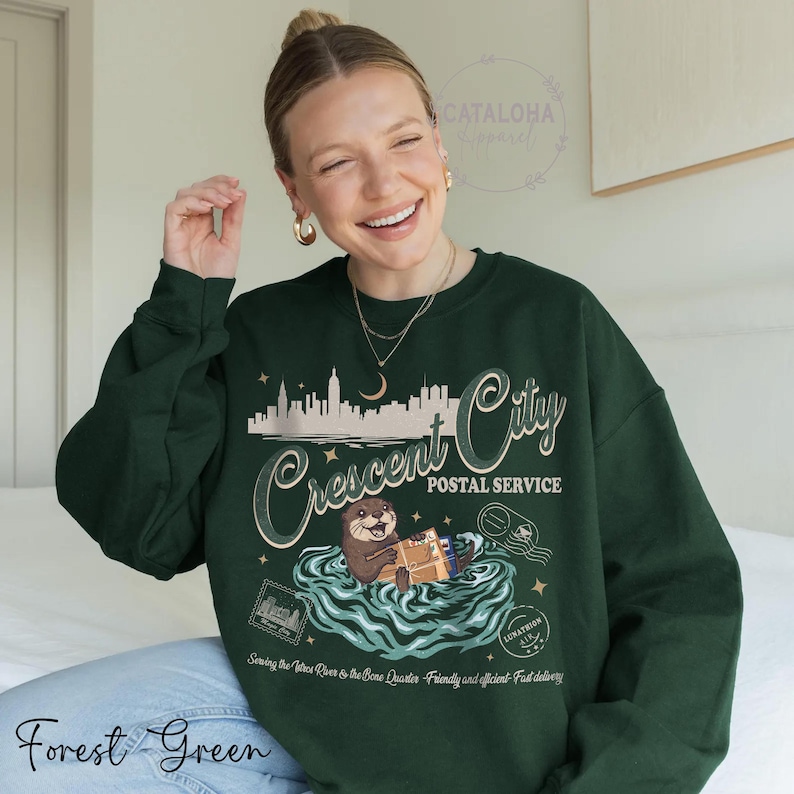 Op de afbeelding: Bost groene sweatshirt met een afbeelding van een otter die post bezorgt voor Crescent City Postal Service. Het ontwerp bevat een stadsgezicht, water en de tekst "Serving the three River to the Bone Quarter. Friendly and efficient. Fast delivery."