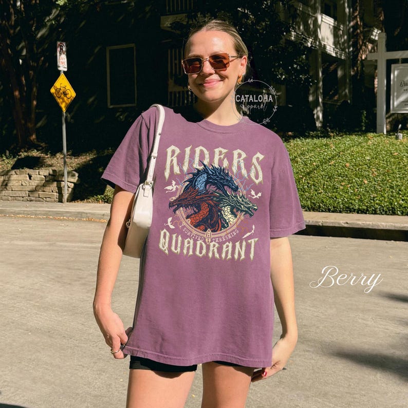 May include: Berry-colored t-shirt with a graphic featuring three dragons and the words "Riders Quadrant". The design includes the text "Survive, Thriving, Crushing". The shirt is a relaxed fit.