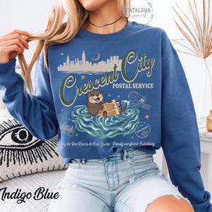Op de afbeelding: Indigo blauwe sweatshirt met een afbeelding van een otter die pakketten in het water bezorgt. De tekst luidt "Crescent City Postal Service" met een stadsbeeld en maanontwerp. De sweatshirt heeft lange mouwen en een ronde hals.