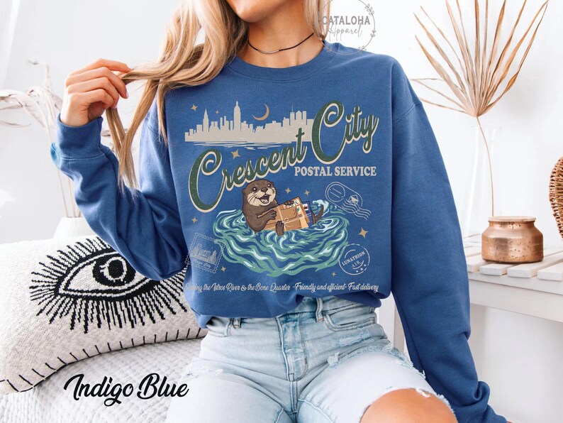 Op de afbeelding: Indigo blauwe sweatshirt met een afbeelding van een otter die pakketten in het water bezorgt. De tekst luidt "Crescent City Postal Service" met een stadsbeeld en maanontwerp. De sweatshirt heeft lange mouwen en een ronde hals.