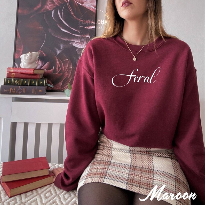 K&ouml;nnte beinhalten: Ein maroonfarbener Pullover mit dem Wort "Feral" in wei&szlig;er Kursivschrift auf der Vorderseite. Der Pullover wird von einer Person getragen, die einen karierten Rock und schwarze Strumpfhosen tr&auml;gt.