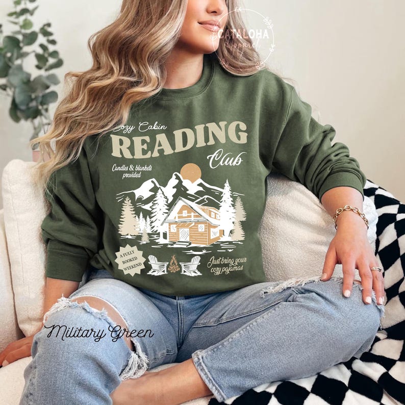 May include: Military green sweatshirt featuring a reading club graphic. The design includes a cabin, mountains, trees, and the text "Cozy Cabin Reading Club." The sweatshirt also has the text "Candles & blanket provided" and "Just bring your cosy pyjamas."
