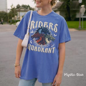 May include: Mystic blue t-shirt with a graphic of three dragons inside a circle, with the words "RIDERS" and "QUADRANT". The shirt has short sleeves and a relaxed fit. The text "Survived Threshing" is also on the shirt.