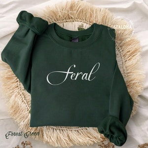 K&ouml;nnte beinhalten: Gr&uuml;n-farbener Sweatshirt mit dem Wort "feral" in wei&szlig;er, kursiv geschriebener Schrift.